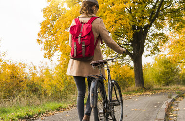 Junge Frau mit Rucksack, die ihr Fahrrad in einer Herbstlandschaft schiebt - UUF006046