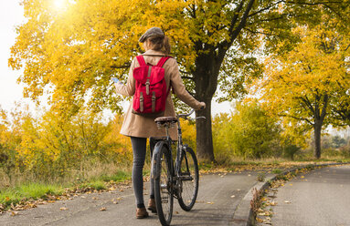 Junge Frau mit Rucksack, die ihr Fahrrad in einer Herbstlandschaft schiebt - UUF006047