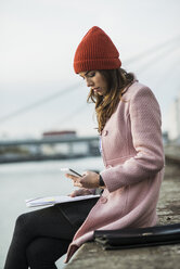 Junge Frau sitzt am Flussufer und schaut auf ihr Handy - UUF006083