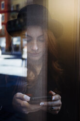 Young woman behind window pane looking at cell phone - GIOF000544