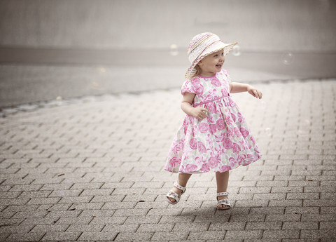 Glückliches blondes kleines Mädchen mit Hut und Sommerkleid mit Blumenmuster tanzt auf dem Bürgersteig, lizenzfreies Stockfoto