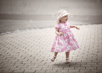 Glückliches blondes kleines Mädchen mit Hut und Sommerkleid mit Blumenmuster tanzt auf dem Bürgersteig - NIF000052