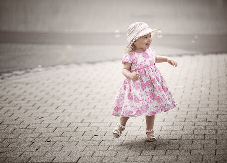 Happy blond little girl wearing hat and summer dress with floral design dancing on pavement - NIF000052