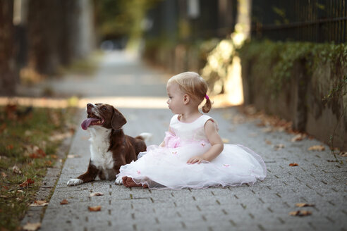 Blond little girl wearing tulle dress sitting besides her dog on pavement - NIF000060
