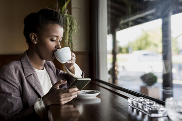 Junge Frau, die in einem Café sitzt, ihr Smartphone benutzt und Kaffee trinkt - MAUF000027