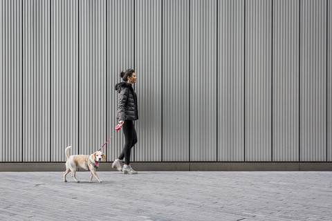 Frau und ihr Hund gehen auf dem Bürgersteig vor einer Metallfassade spazieren, lizenzfreies Stockfoto