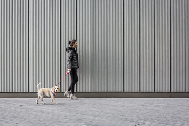 Frau und ihr Hund gehen auf dem Bürgersteig vor einer Metallfassade spazieren - MAUF000052