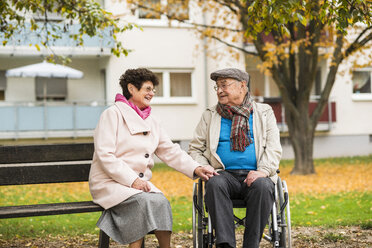 Senior woman sitting on bench next to husband in wheelchair - UUF006118
