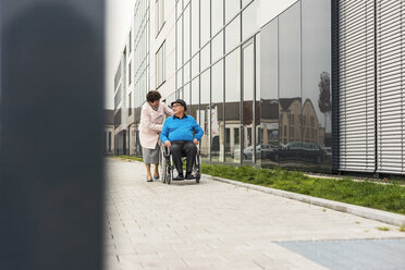 Senior woman pushing husband in wheelchair on pavement - UUF006127