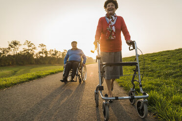 Lächelndes Seniorenpaar mit Rollstuhl und Rollator auf einem Weg - UUF006157