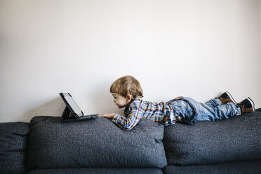 Kleiner Junge liegt auf der Rückenlehne einer Couch und schaut auf ein digitales Tablet - JRFF000200