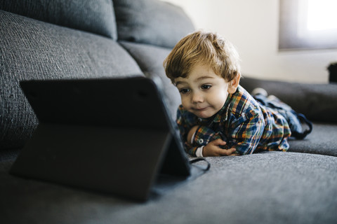 Porträt eines kleinen Jungen, der auf der Couch liegt und auf ein digitales Tablet schaut, lizenzfreies Stockfoto