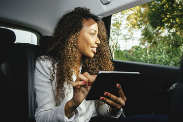Smiling young woman sitting on back seat of a car holding mini tablet - EBSF001135