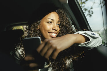 Lächelnde junge Frau sitzt in einem Auto mit Smartphone und schaut durch das Fenster - EBSF001137