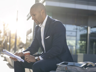 Germany, Cologne, Young businessman sitting on bench, reading magazine - MADF000683