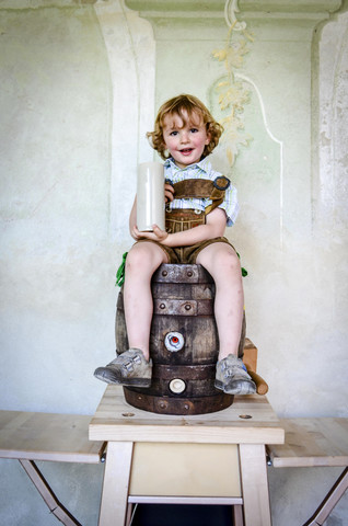 Deutschland, Bayern, Porträt eines lächelnden kleinen Jungen, der auf einem Holzfass sitzt und einen Bierkrug hält, lizenzfreies Stockfoto