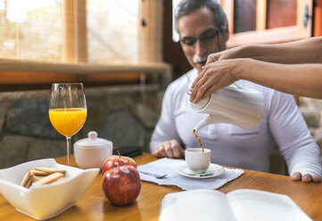 Mann sitzt beim Frühstück, während eine Frau ihm Kaffee in die Tasse gießt - MGOF001090