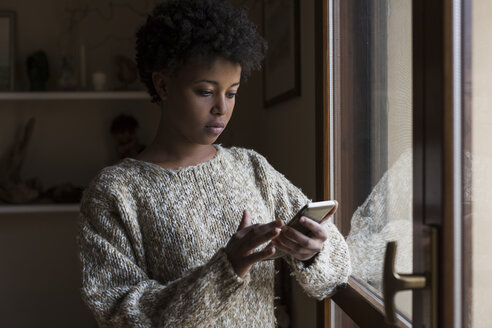 Junge Frau am Fenster, die auf ihr Smartphone schaut - MAUF000111