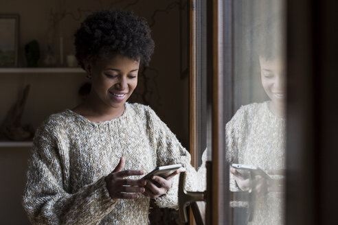 Lächelnde junge Frau am Fenster, die auf ihr Smartphone schaut - MAUF000112