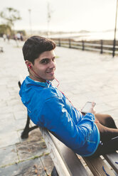 Smiling athlete sitting on bench after training listening to music from smartphone - RAEF000687