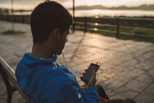 Sportler sitzt nach dem Training auf einer Bank und hört bei Sonnenuntergang Musik vom Smartphone - RAEF000690