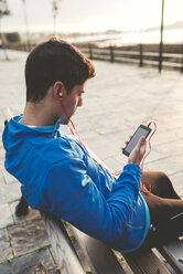 Sportler sitzt nach dem Training auf der Bank und hört Musik vom Smartphone - RAEF000691