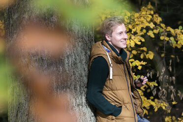 Smiling young man leaning at tree in autumn - GDF000920