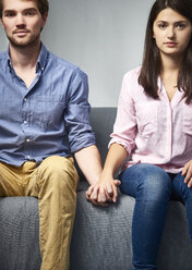 Ernstes junges Paar sitzt auf einer Couch und hält sich die Hände - DISF002263