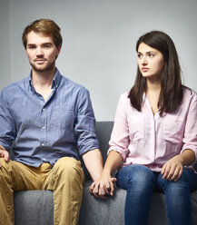 Ernstes junges Paar sitzt auf einer Couch und hält sich die Hände - DISF002264