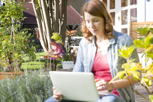 Junge Frau mit Laptop im Garten mit Mutter und Tochter im Hintergrund - FKF001636