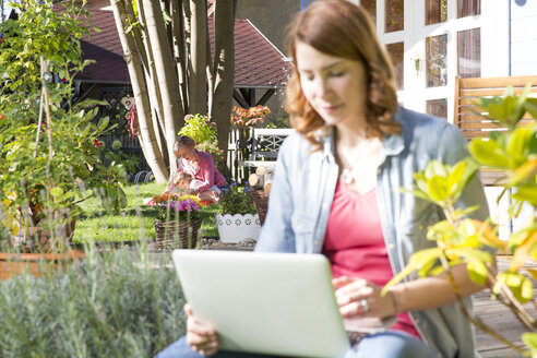 Young woman using laptop in garden with mother and daughter in background - FKF001636