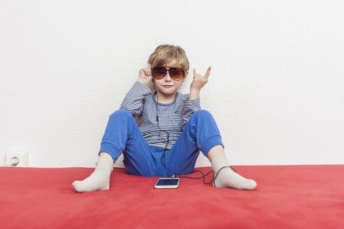 Kleiner Junge mit Sonnenbrille sitzt auf dem Bett und hört Musik mit Smartphone und Kopfhörern - MMFF001276