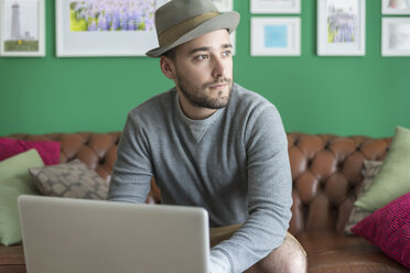Porträt eines bärtigen jungen Mannes mit Hut, der mit einem Laptop auf einer Couch sitzt - PAF001494