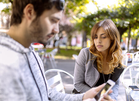 Porträt einer jungen Frau, die ihren Freund beim Benutzen seines Smartphones beobachtet - MGOF001122