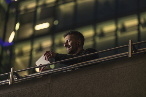 Deutschland, Berlin, lächelnder Mann mit Blick auf Mini-Tablet vor beleuchtetem Bürogebäude - MMFF001279