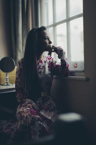 Junge Frau in geblümtem Kleid schaut aus dem Fenster, lizenzfreies Stockfoto