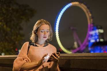 Großbritannien, London, junge Frau schaut auf ihr Smartphone mit London Eye im Hintergrund - MAUF000136