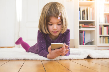 Kleines Mädchen liegt auf einer Decke auf dem Boden und schaut auf ein Smartphone - LVF004247