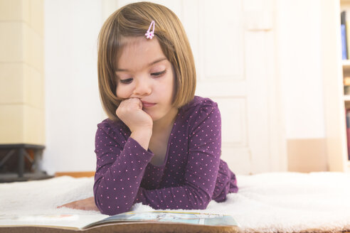 Pensive little girl lying on blanket on the floor watching picture book - LVF004251