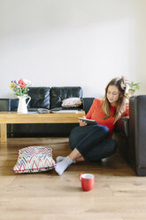 Junge Frau sitzt auf dem Boden ihres Wohnzimmers und schaut auf ihr digitales Tablet - BOYF000018