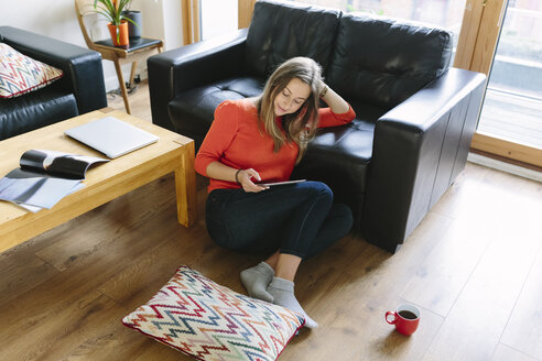 Junge Frau sitzt auf dem Boden ihres Wohnzimmers und schaut auf ihr digitales Tablet - BOYF000019