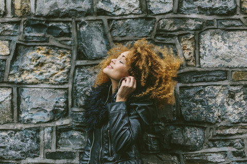 Frau mit Afro hört Musik mit Kopfhörern vor einer Natursteinmauer, lizenzfreies Stockfoto