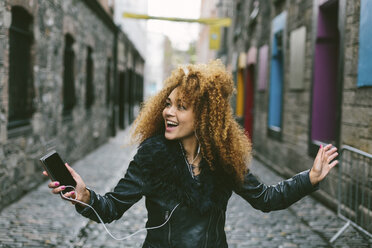 Irland, Dublin, fröhliche Frau mit Afro, die mit Smartphone und Kopfhörern Musik hört - BOYF000037
