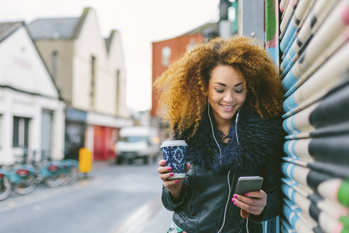 Irland, Dublin, lächelnde Frau mit Kaffee zum Mitnehmen, die mit Smartphone und Kopfhörern Musik hört - BOYF000042