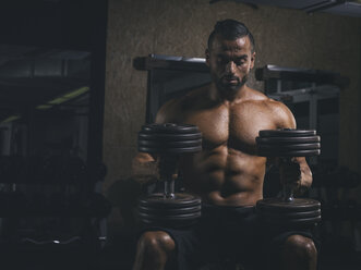 Bodybuilder resting with heavy dumbells on his knees in gym - MADF000770