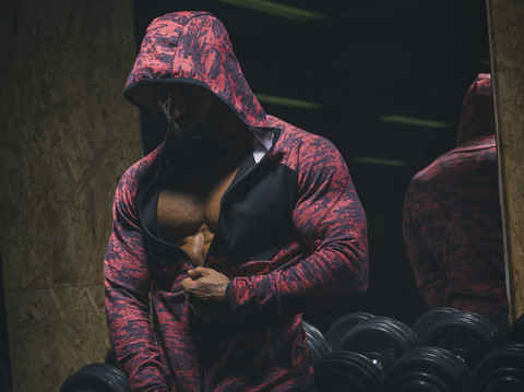 Bodybuilder, der seinen Kapuzenpullover mit Reißverschluss vor einem Hantelständer in einem Fitnessstudio schließt, lizenzfreies Stockfoto