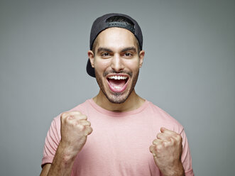 Portrait of man with baseball cap screaming for joy in front of grey background - RH001077