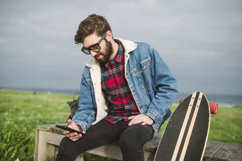 Junger lässiger Mann sitzt mit seinem Longboard in einem Park, er schaut auf sein Smartphone - RAEF000711