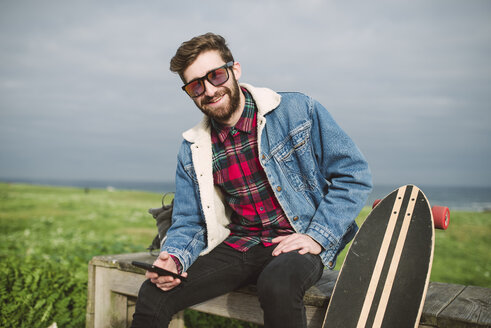 Junger lässiger Mann sitzt mit seinem Longboard in einem Park, er lächelt und schaut in die Kamera - RAEF000712