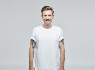 Porträt eines lächelnden Mannes mit Tatoos auf den Armen, der ein weißes T-Shirt vor einem grauen Hintergrund trägt - RHF001090